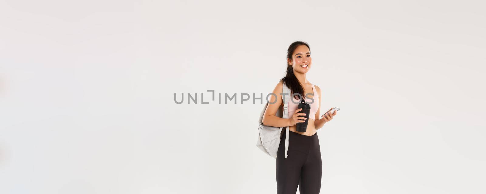 Full length of smiling healthy, slim asian girl going fitness training, female athelte carry backpack with workout equipment and water bottle, using mobile phone sports application, white background by Benzoix