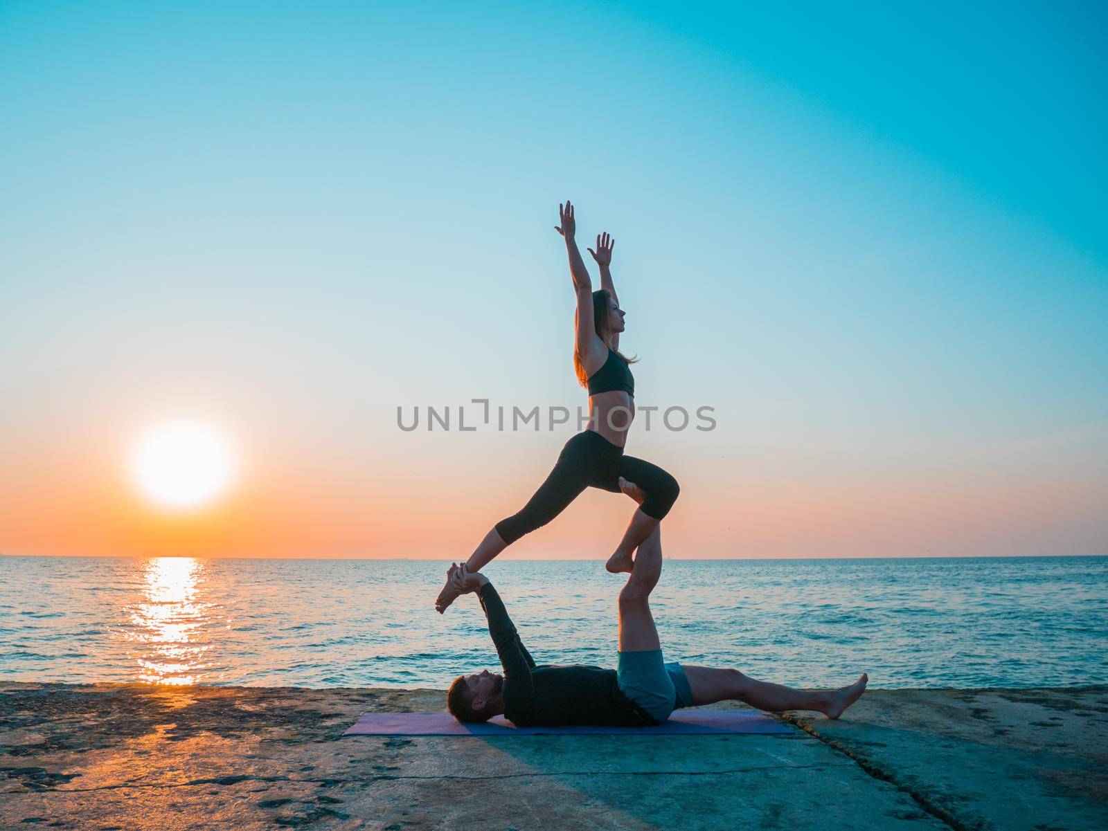 Fit young couple doing acro-yoga at sea beach. Man lying on concrete plates and balancing woman on his feet. Beautiful pair practicing yoga together. by kristina_kokhanova