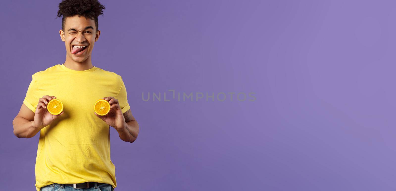 Holidays, vitamins and vacation concept. Portrait of sassy and cheeky funny young playful guy, fool around with fruit, holding pieces of oranges like women breast, show tongue smiling by Benzoix