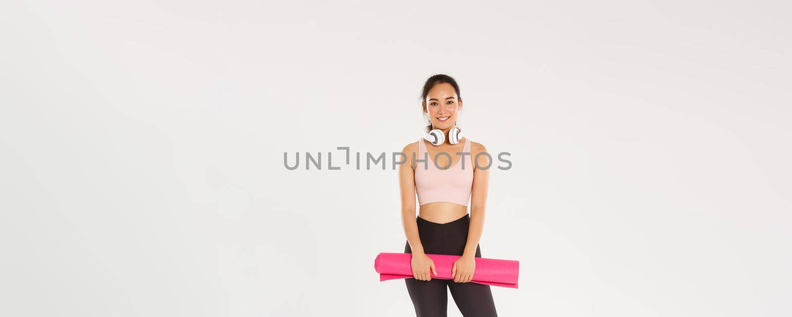 Full length of smiling slim asian girl with rubber mat for fitness exercises, headphones, looking at camera while waiting for training coach starting workout session in gym, white background.