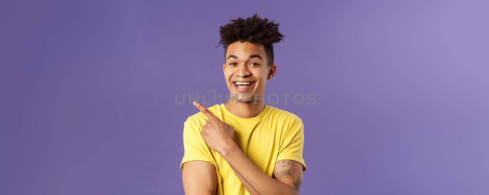 Close-up portrait of cheerful, attractive young male student showing promo, pointing at upper left corner, smiling pleased, recommend click link, visit page or download app, purple background.