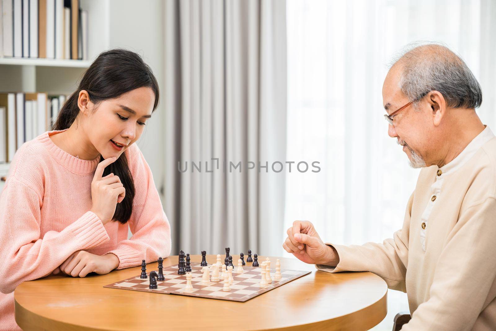 Beautiful young smile woman having fun sitting playing chess game with senior elderly at home by Sorapop