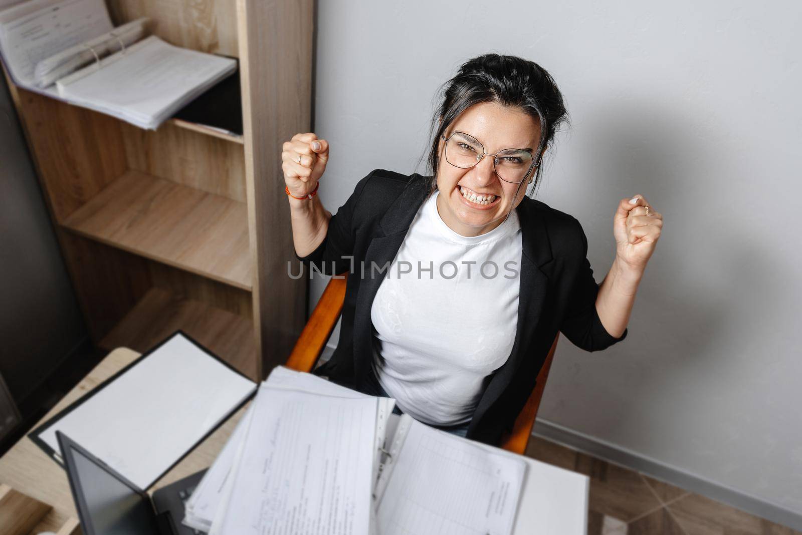 An angry office employee. The girl manager working with papers at her laptop is very angry and looks at the camera with anger. Stress at work, feeling unwell.
