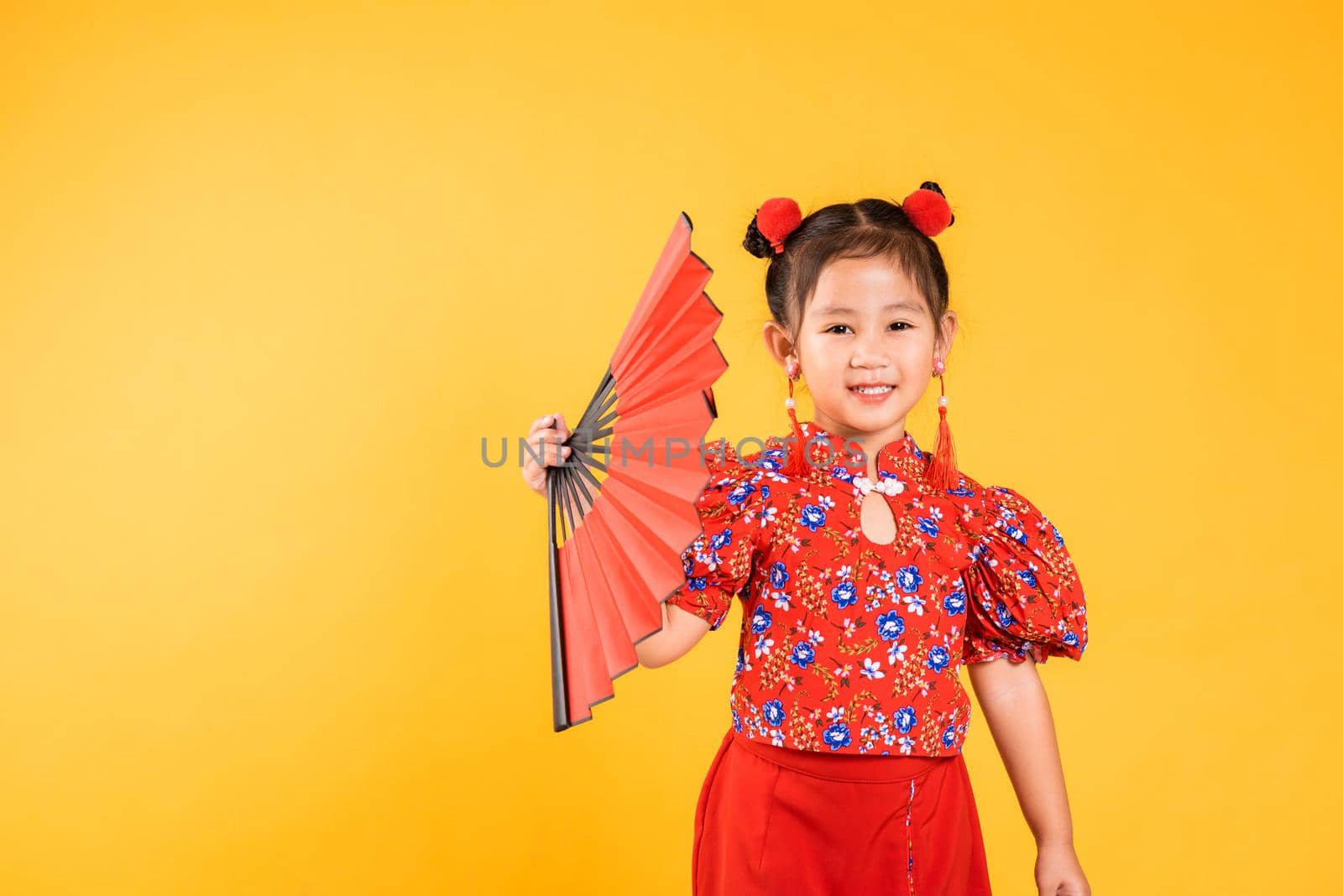 Chinese New Year. Happy Asian Chinese little girl smile wearing red cheongsam holding fan, Portrait children in traditional dress, studio short isolated on yellow background