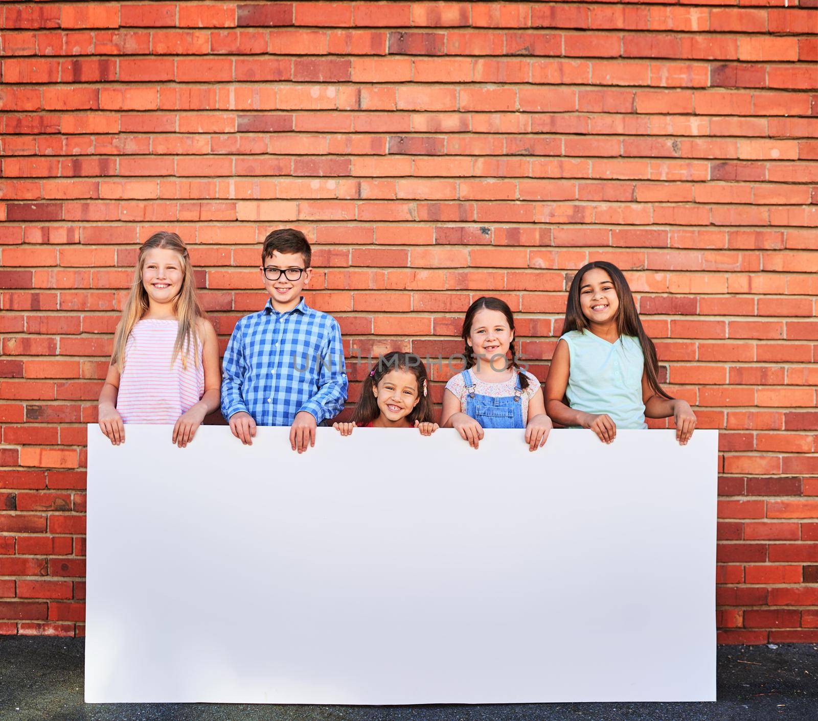 Well use our cuteness to help promote your message. Portrait of a group of young children holding a blank sign against a brick wall. by YuriArcurs