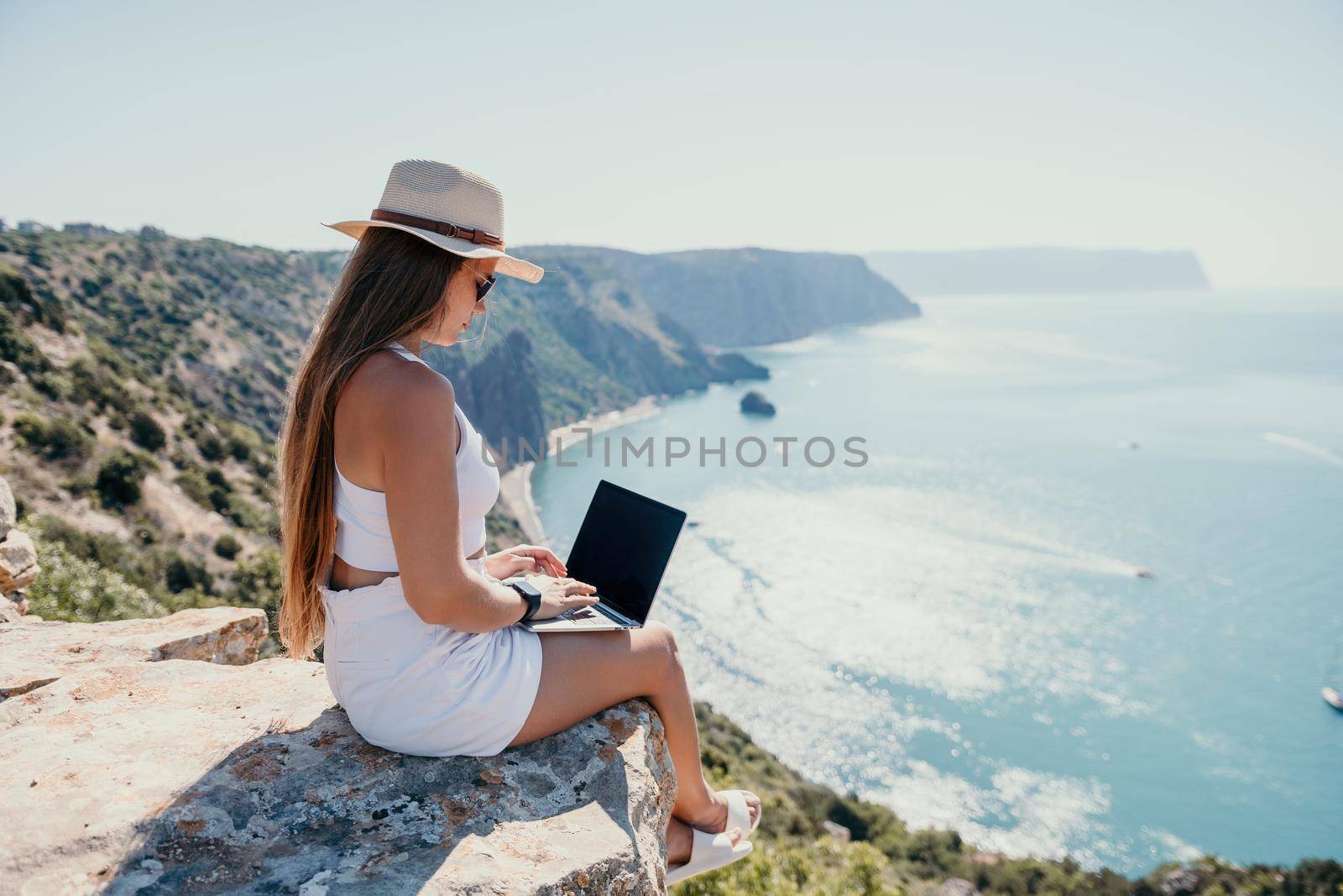 Successful business woman in yellow hat working on laptop by the sea. Pretty lady typing on computer at summer day outdoors. Freelance, travel and holidays concept.