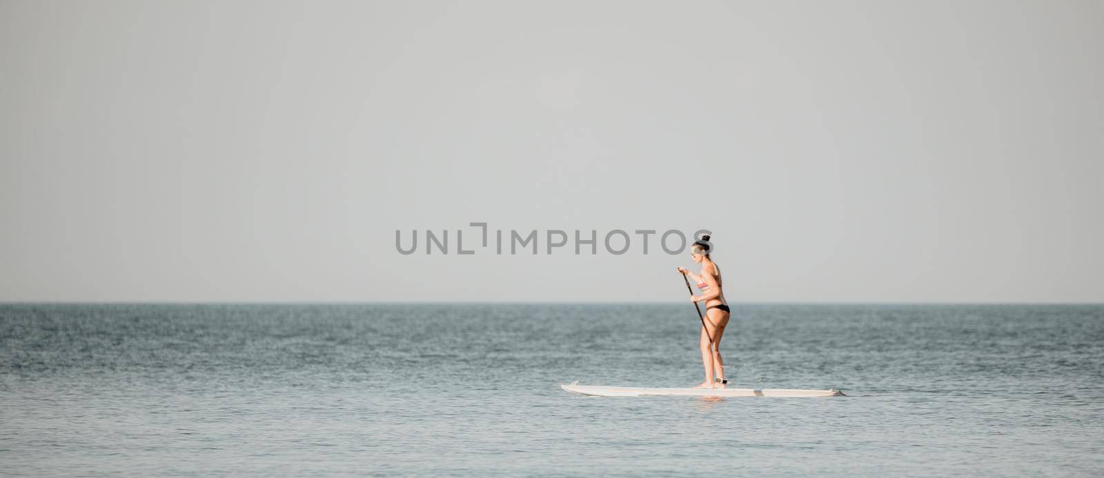 Silhouette of woman standing, surfing on SUP board, confident paddling through water surface. Idyllic sunset or sunrise. Sports active lifestyle at sea or river.