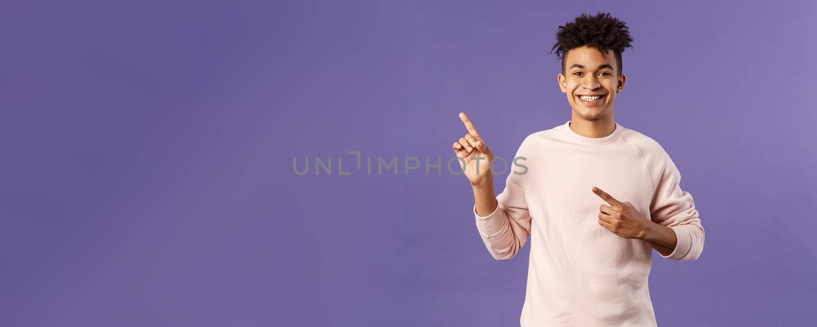 Portrait of cheerful, healthy smiling man pointing fingers upper left corner, recommending buy product, shop online, order delivery, advertisement and people concept, purple background.