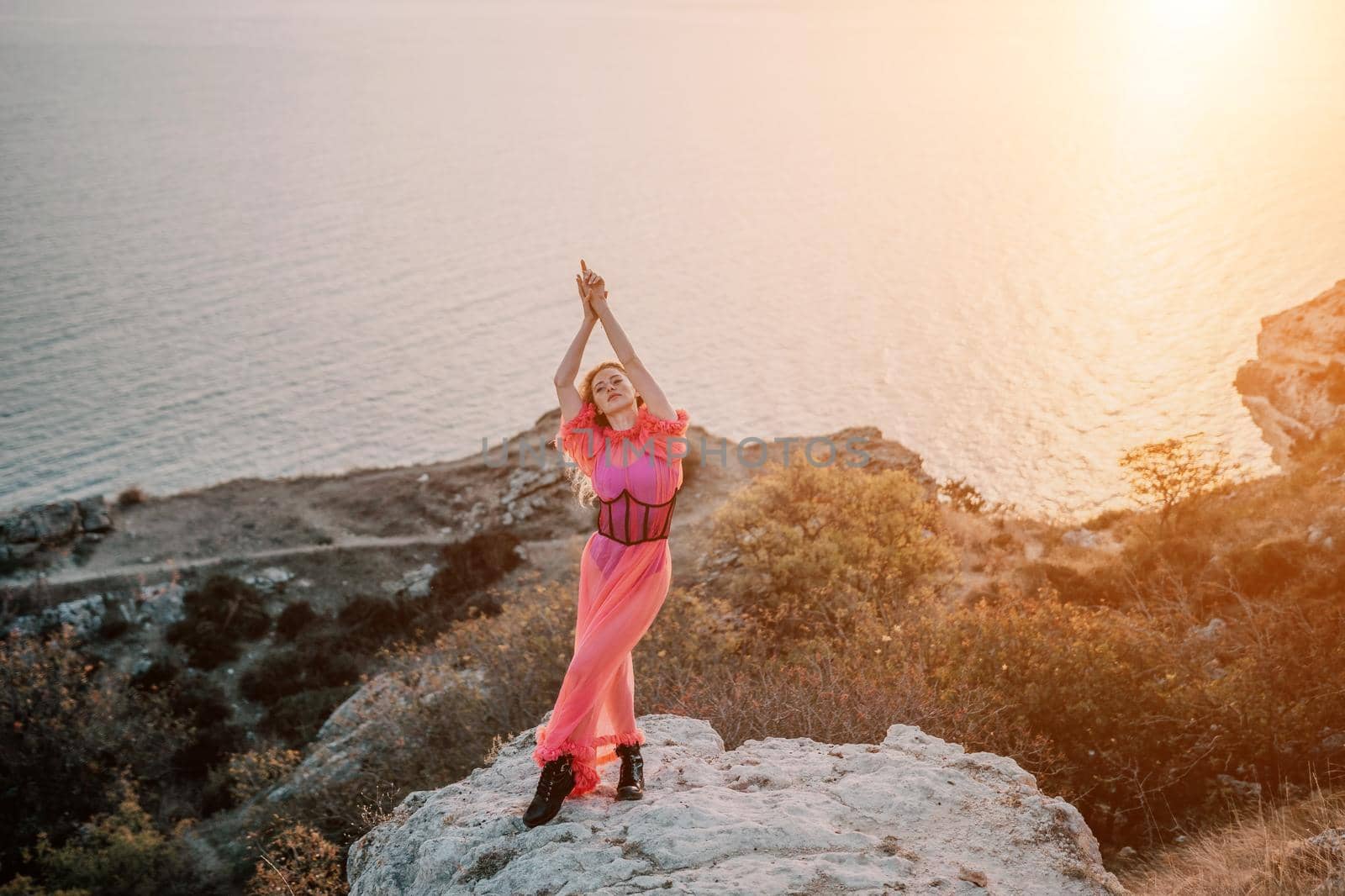 Beautiful young caucasian woman with curly blond hair and freckles. Cute redhead woman portrait in a pink long dress posing on a volcanic rock high above the sea during sunset. by panophotograph