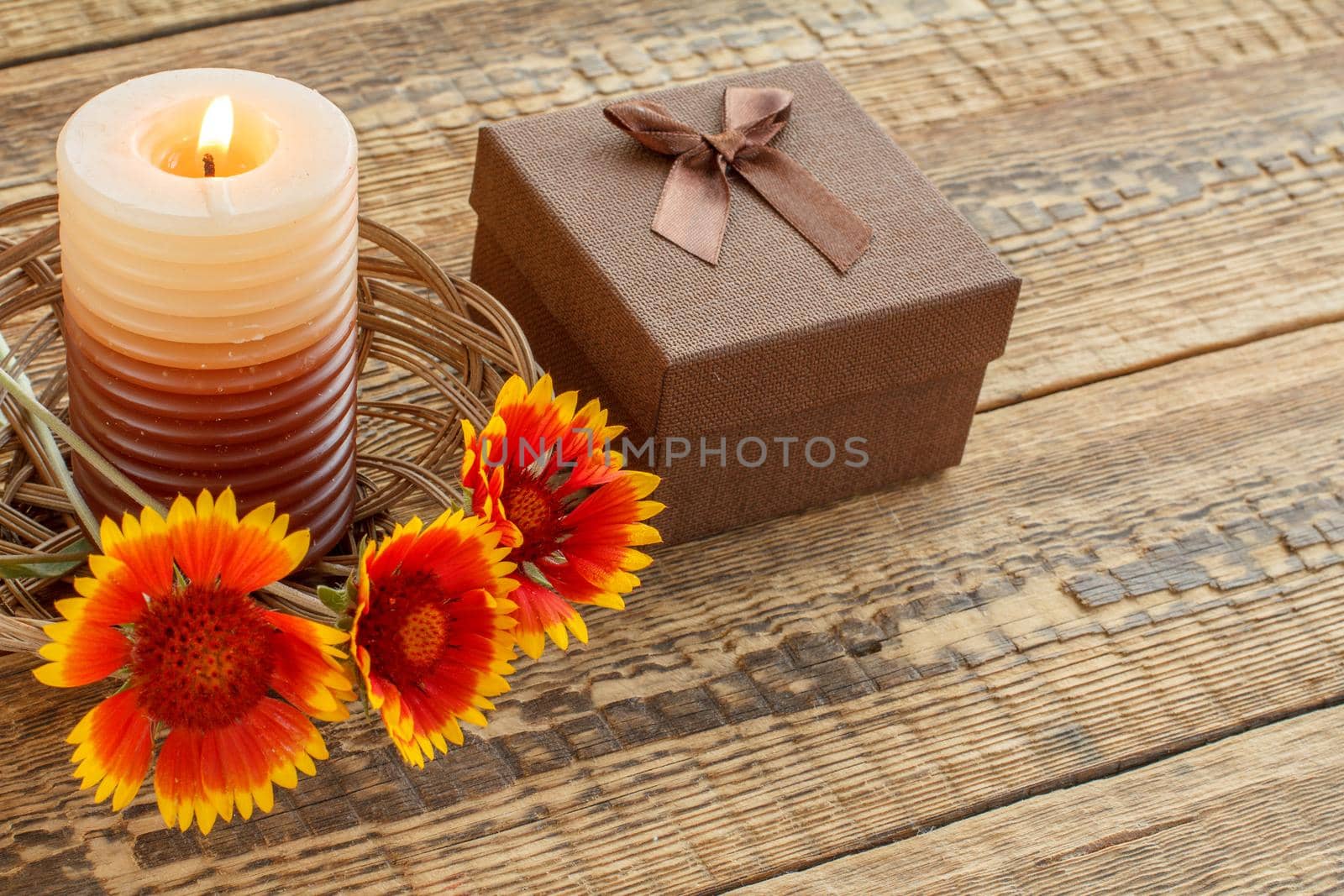 Burning candle, brown gift box wrapped in kraft paper with red ribbon and flowers on wooden boards. Top view. Holiday concept.