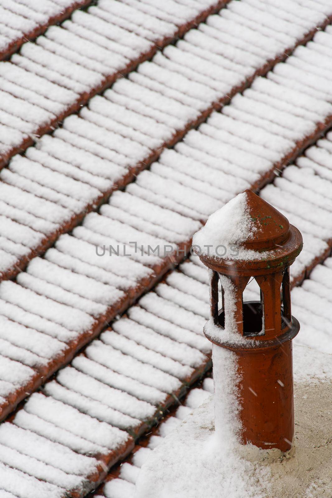 Elevated view of snow covered roof with red tiles and chimney pipe close up during a winter day.