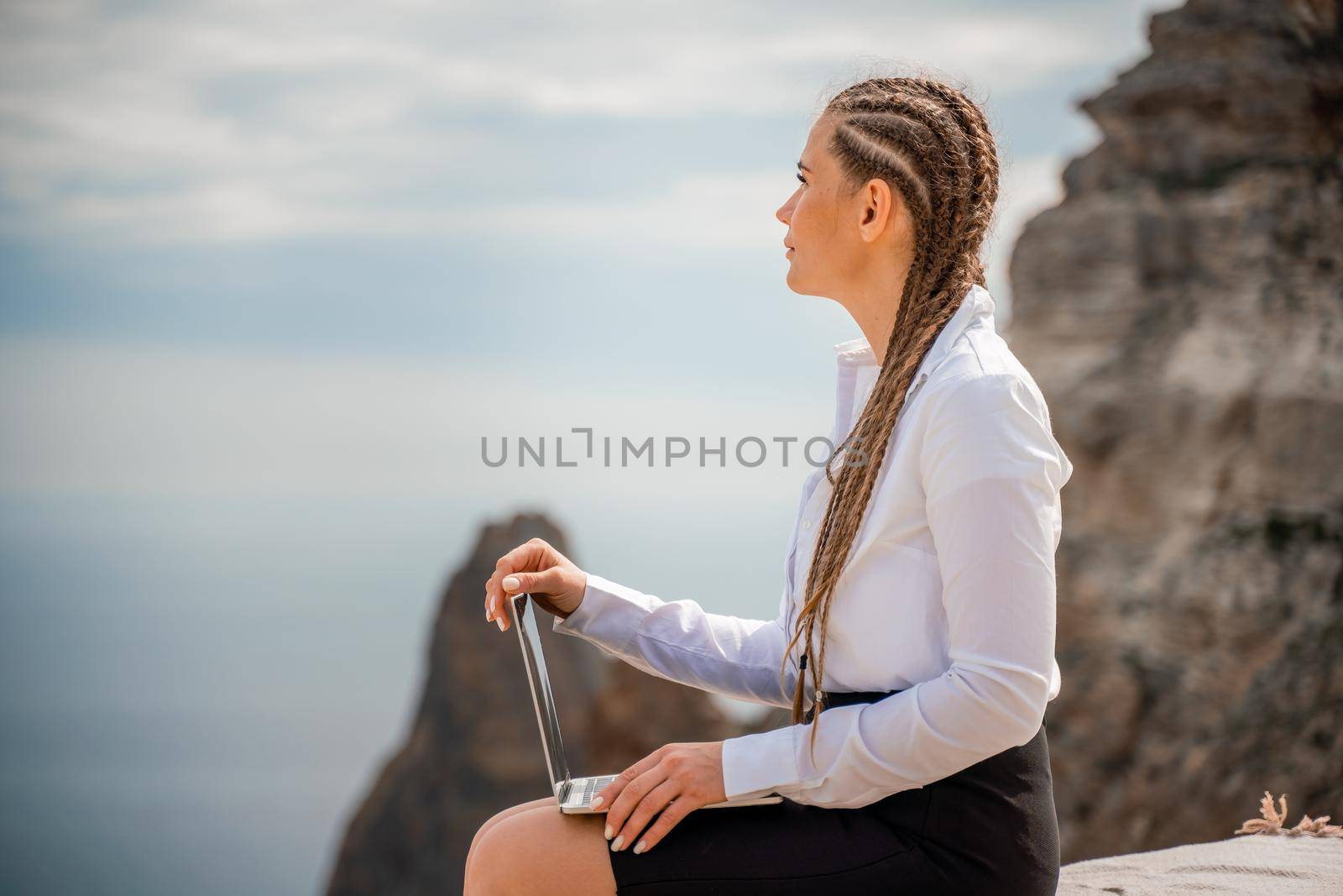 A woman is sitting and typing on a macbook keyboard on a terrace with a beautiful sea view. Wearing a white blouse and black skirt. Freelance travel and vacation concept, digital nomad