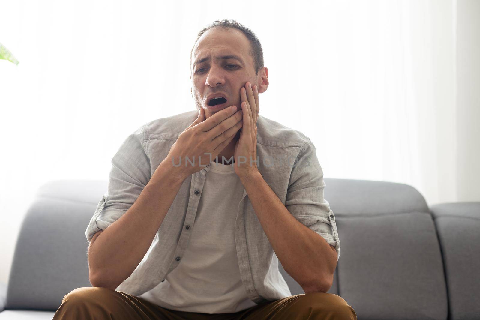 Tooth ache concept. Young man feeling pain, holding his cheek with both hands, suffering from bad toothache, having painful expression by Andelov13