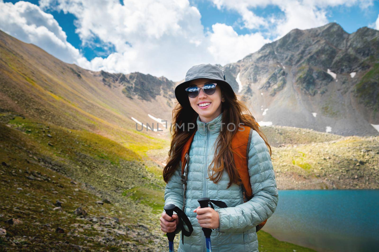 Smiling, positive girl with disheveled hair against the background of a blue mountain lake, with trekking poles in her hands and a backpack, a tourist on a hike.