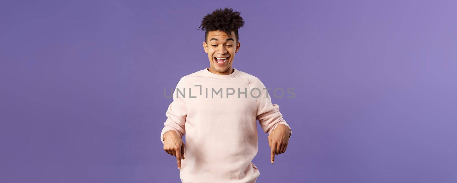 Joyful good-looking hipster guy with dreads rejoicing over good news, best deal ever in online store, favorite game finally in stock, pointing fingers down and rejoicing, purple background.