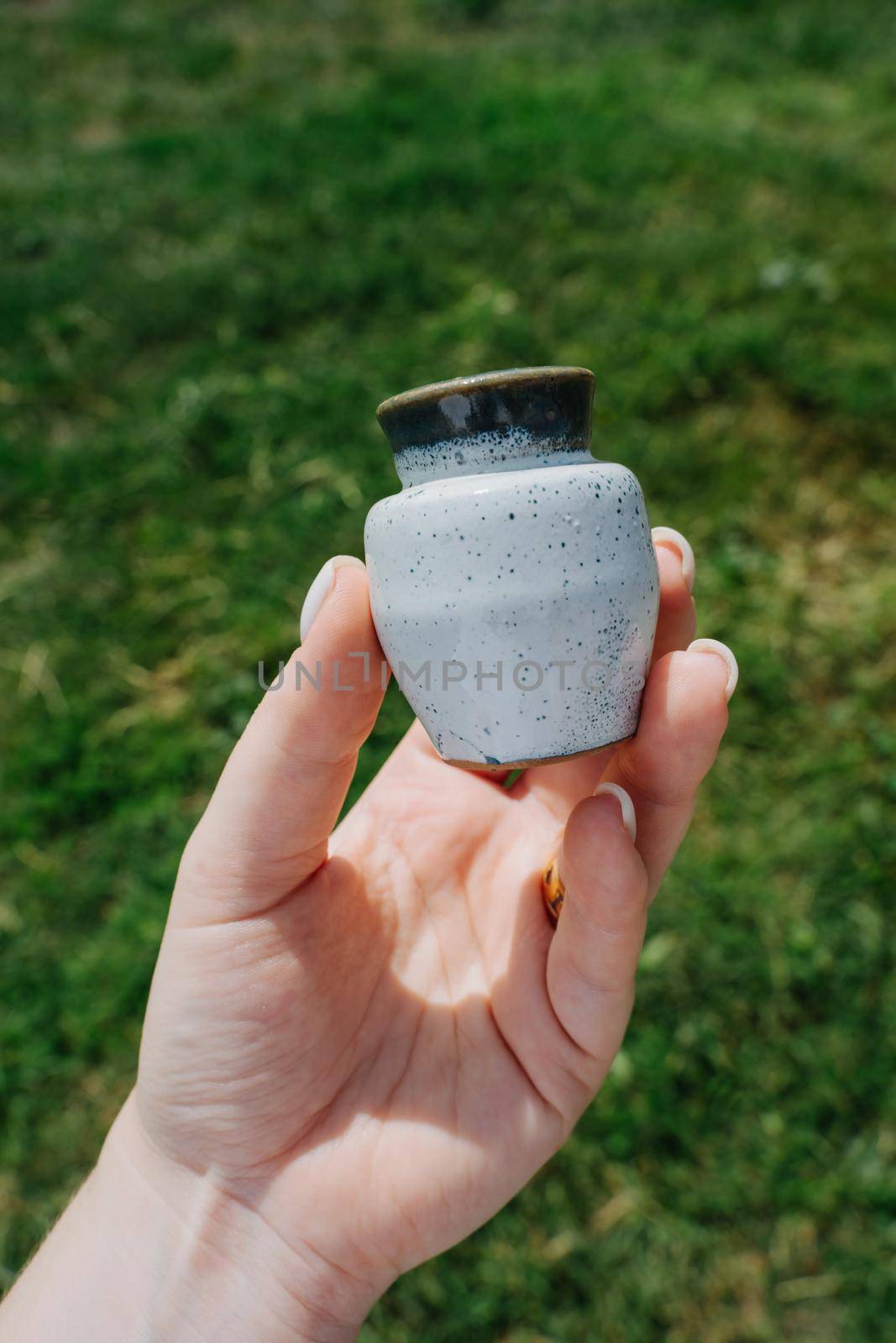Small ceramic jug. The girl's hand holds a small clay jug of light gray color. Juicy green grass on the background. Vertical photo.