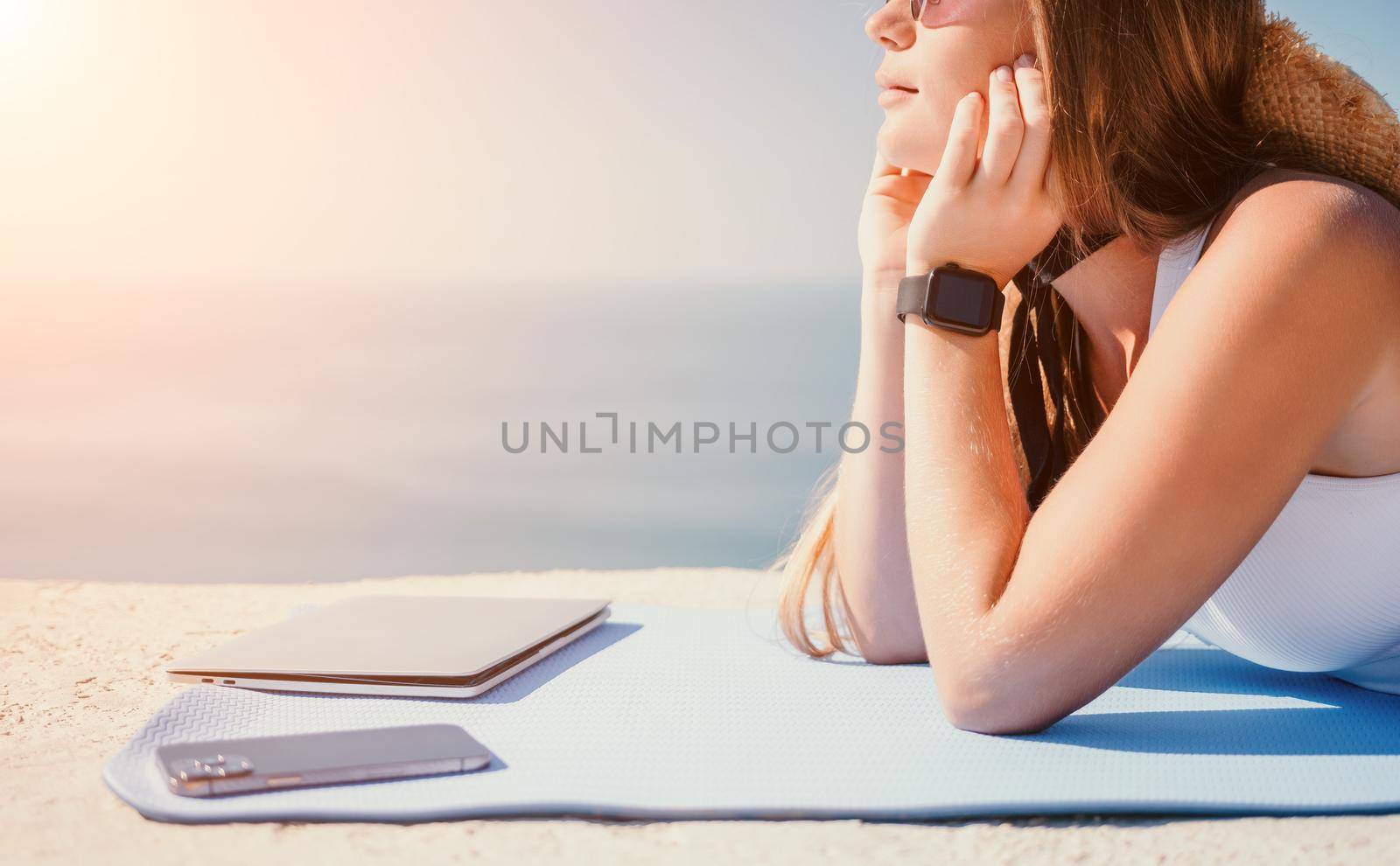 Successful business woman in yellow hat working on laptop by the sea. Pretty lady typing on computer at summer day outdoors. Freelance, travel and holidays concept.