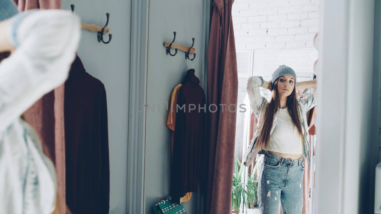 Mirror shot of young girl trying on clothes in fitting room. She is wearing denim jacket, jeans and hat, smoothing her hair, moving and looking in mirror to check image. by silverkblack