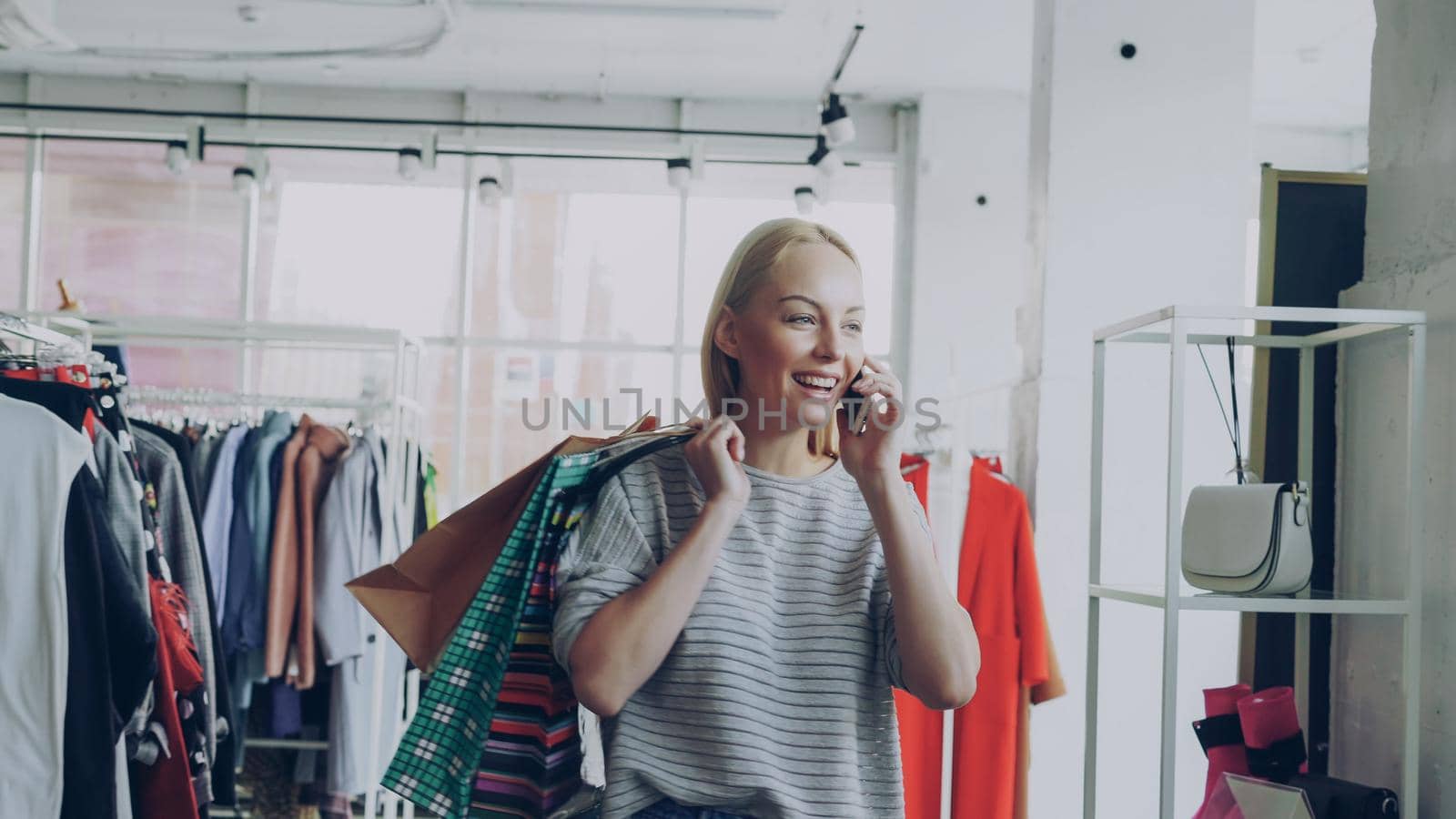 Attractive blond woman is walking between shelves and rails in large store and talking on mobile phone. She is carrying bags, smiling and looking at trendy clothes around her. by silverkblack