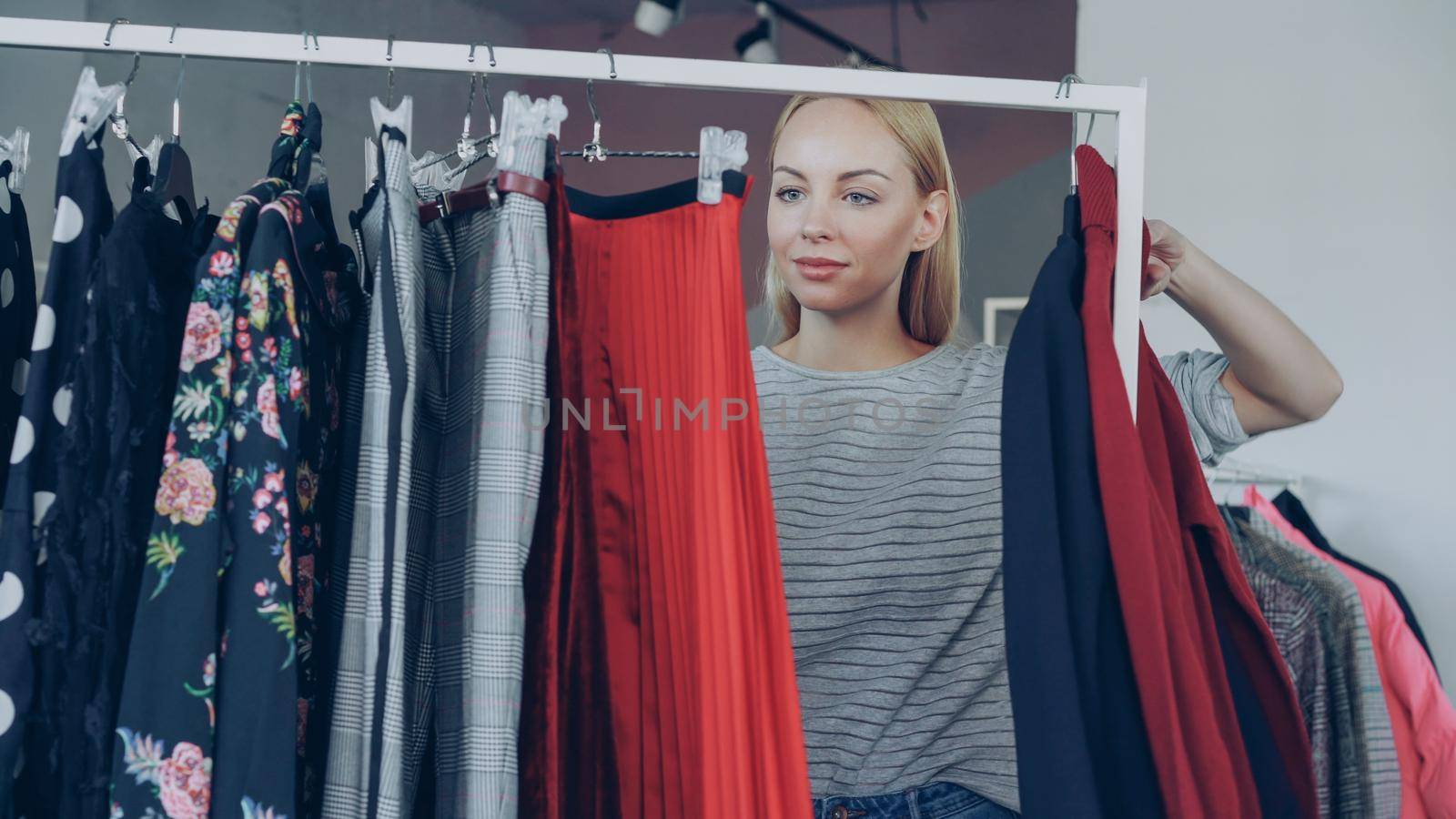 Blonde young lady shopping for clothes, looking through colourful garments on rails, touching and moving them. Women's skirts, jumpers and jackets in the foreground.