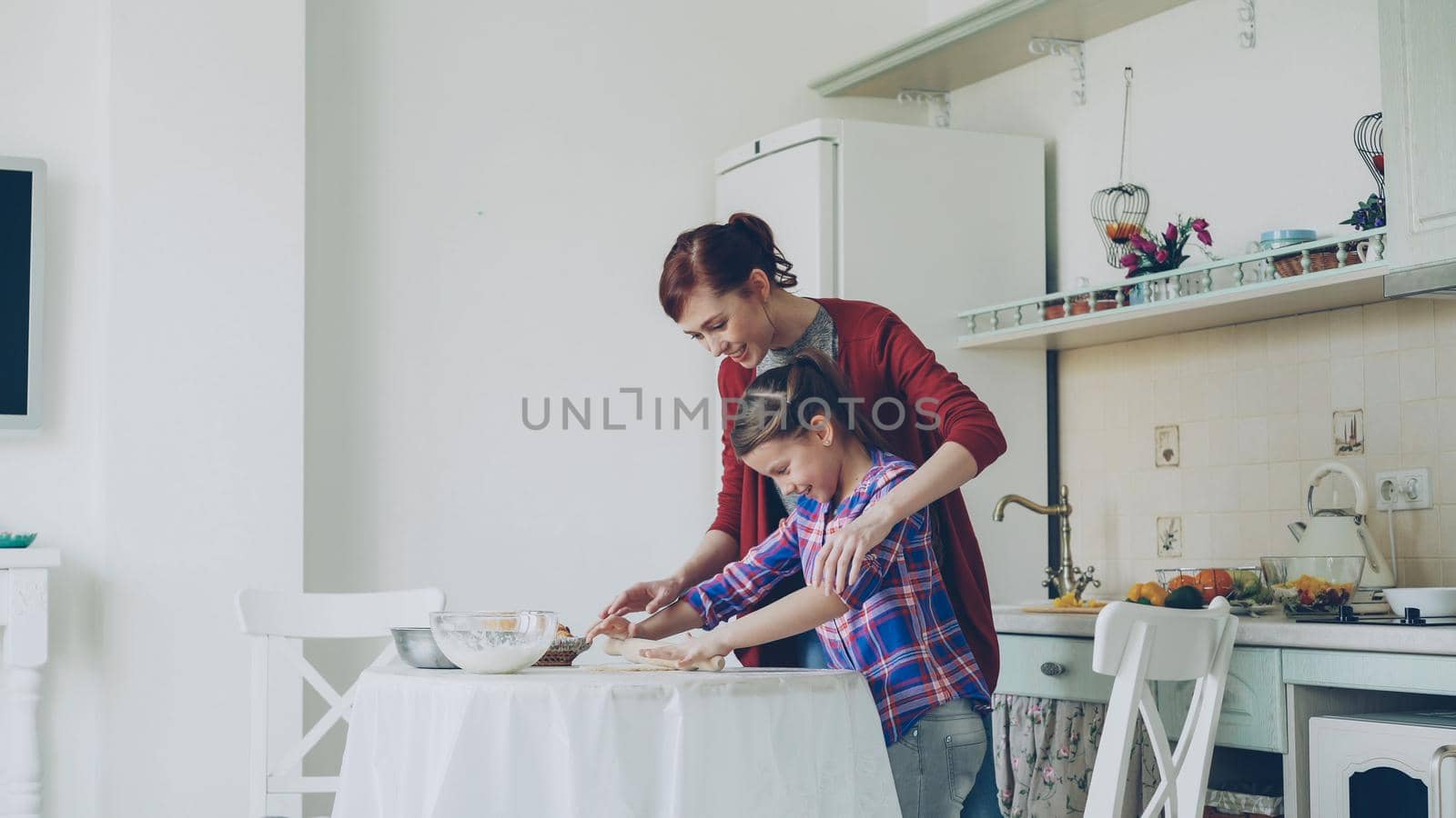 Young attractive mother teaching her little cute daughter rolling dough while cooking together in the kitchen at home on holidays. Family, food and people concept