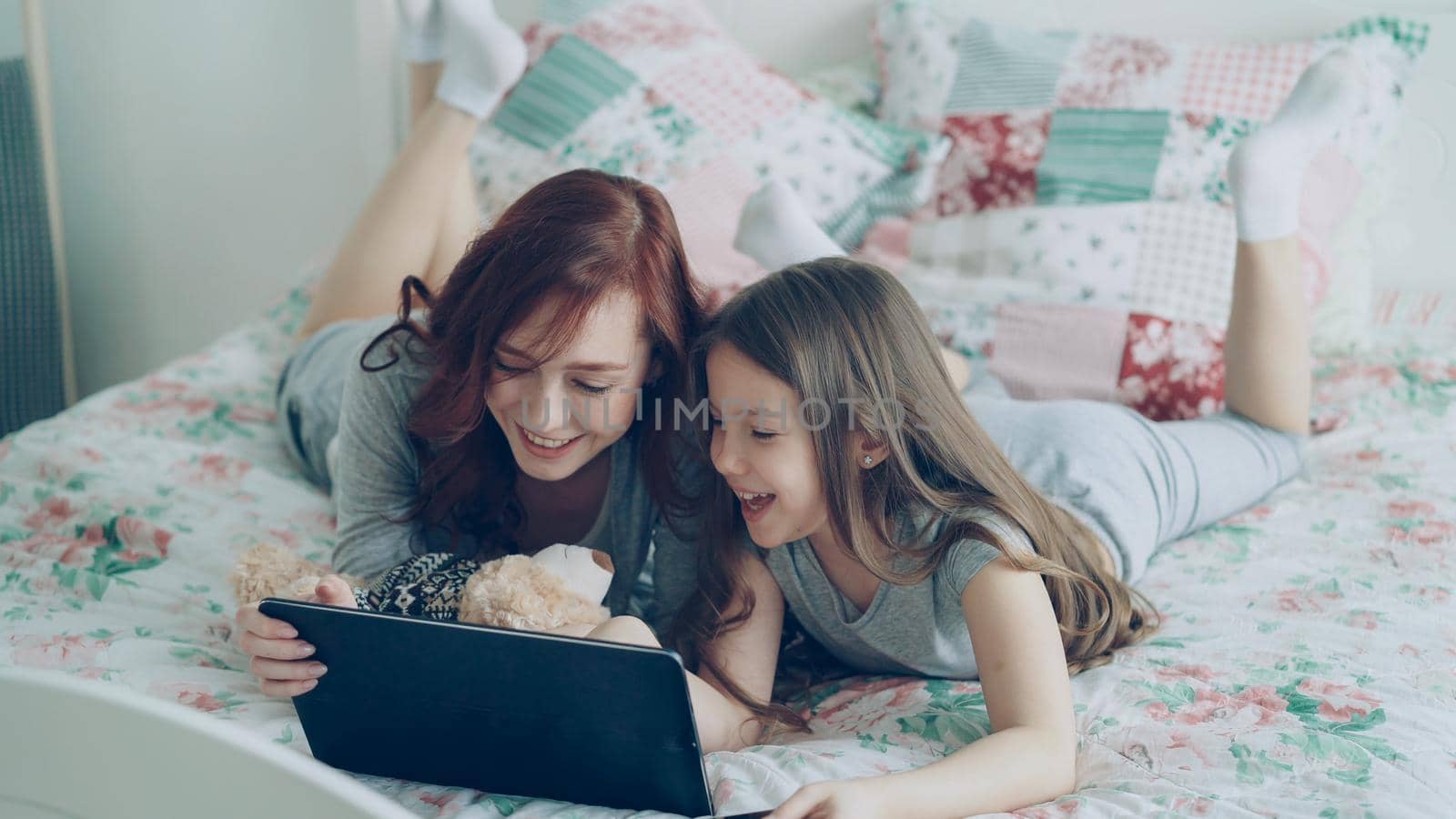 Attractive young mother and her cute daughter in pajamas laughing and looking in digital tablet while lying on bed at home in the morning