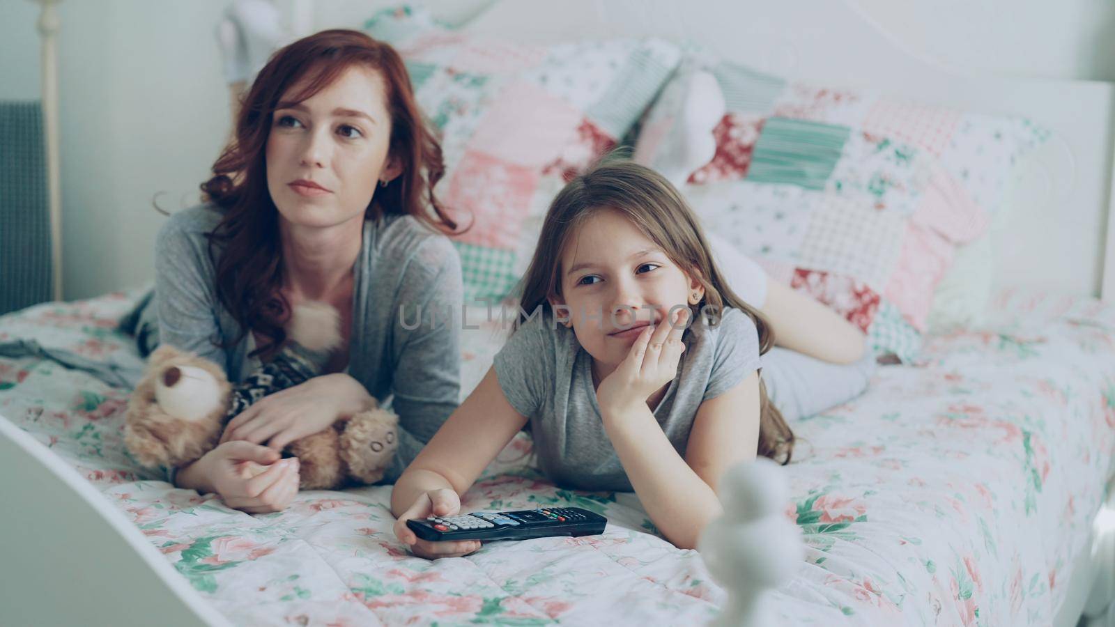 Cheerful happy mother with little daughter watching cartoon movie on TV using remote and smiling while lying on bed at home in cozy bedroom