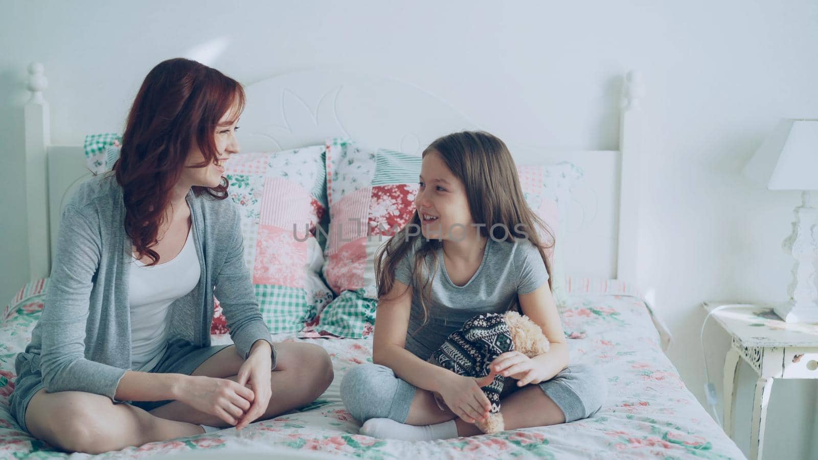 Young mother and little cheerful daughter talking and sharing secrets while sitting on bed at home in bedroom. Relationship and family concept