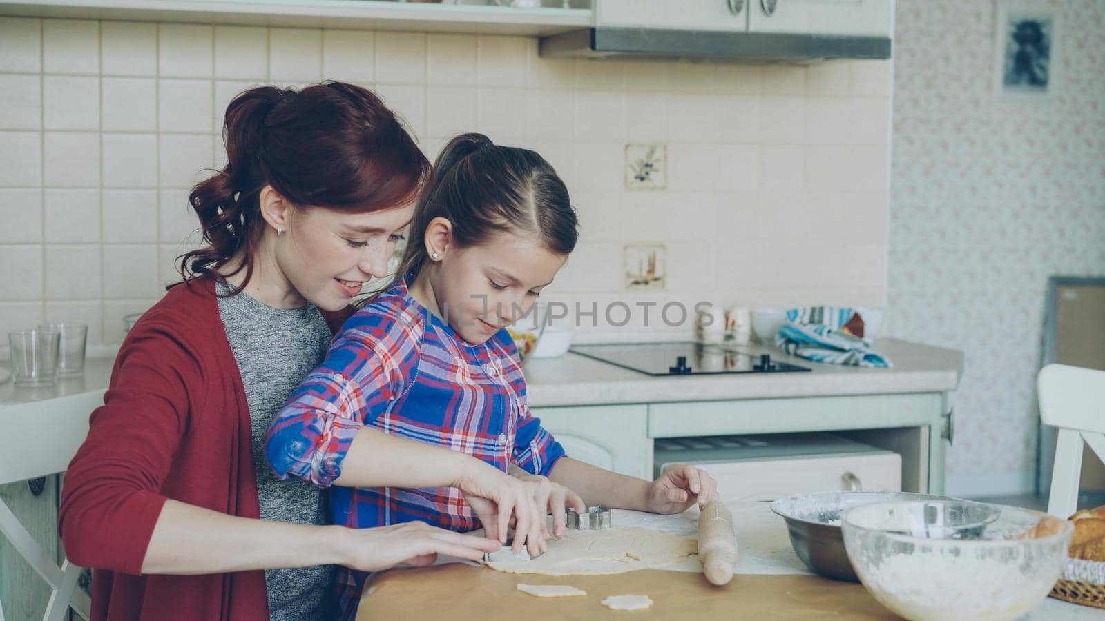 Smiling mother and cute funny daughter making cookies together using bakery forms while sitting in modern kitchen at home. Family, food and people concept