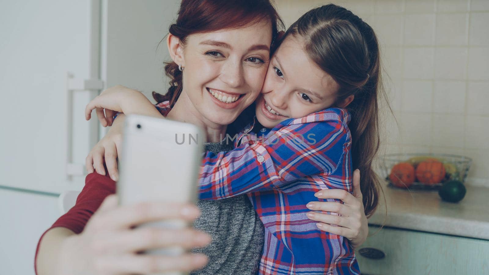 Smiling mother together with funny playful daughter making selfie photo with smartphone camera at home in kitchen. Family, cooking, and people concept