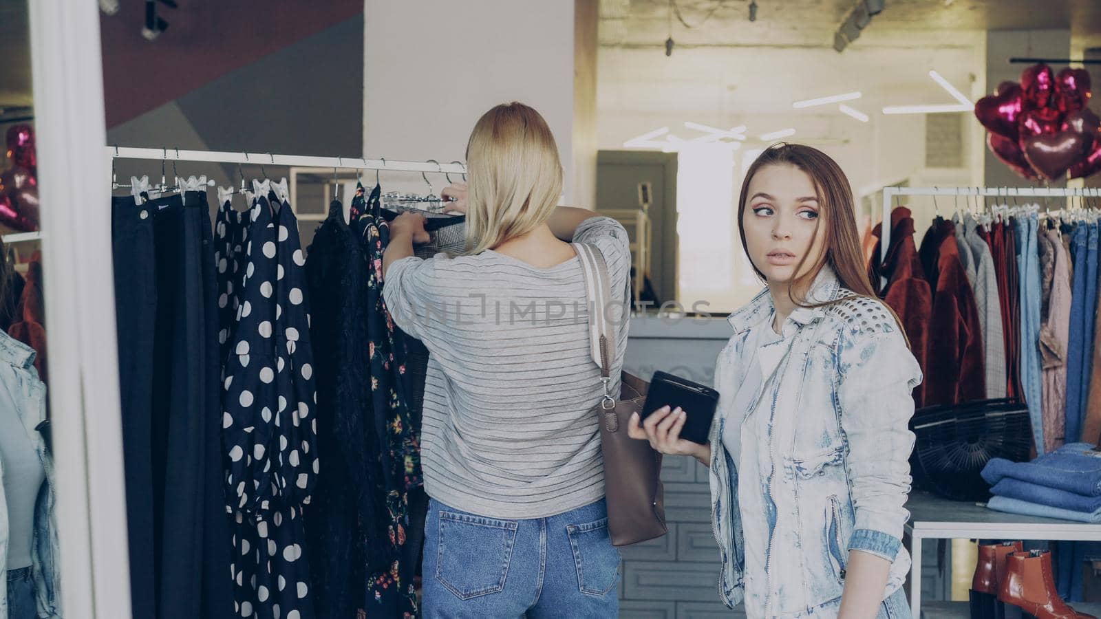 Young woman stealing wallet from female customer choosing clothes in shop. Thief looking around carefully and walking away. Fine boutique with clothing, bags and shoes in background