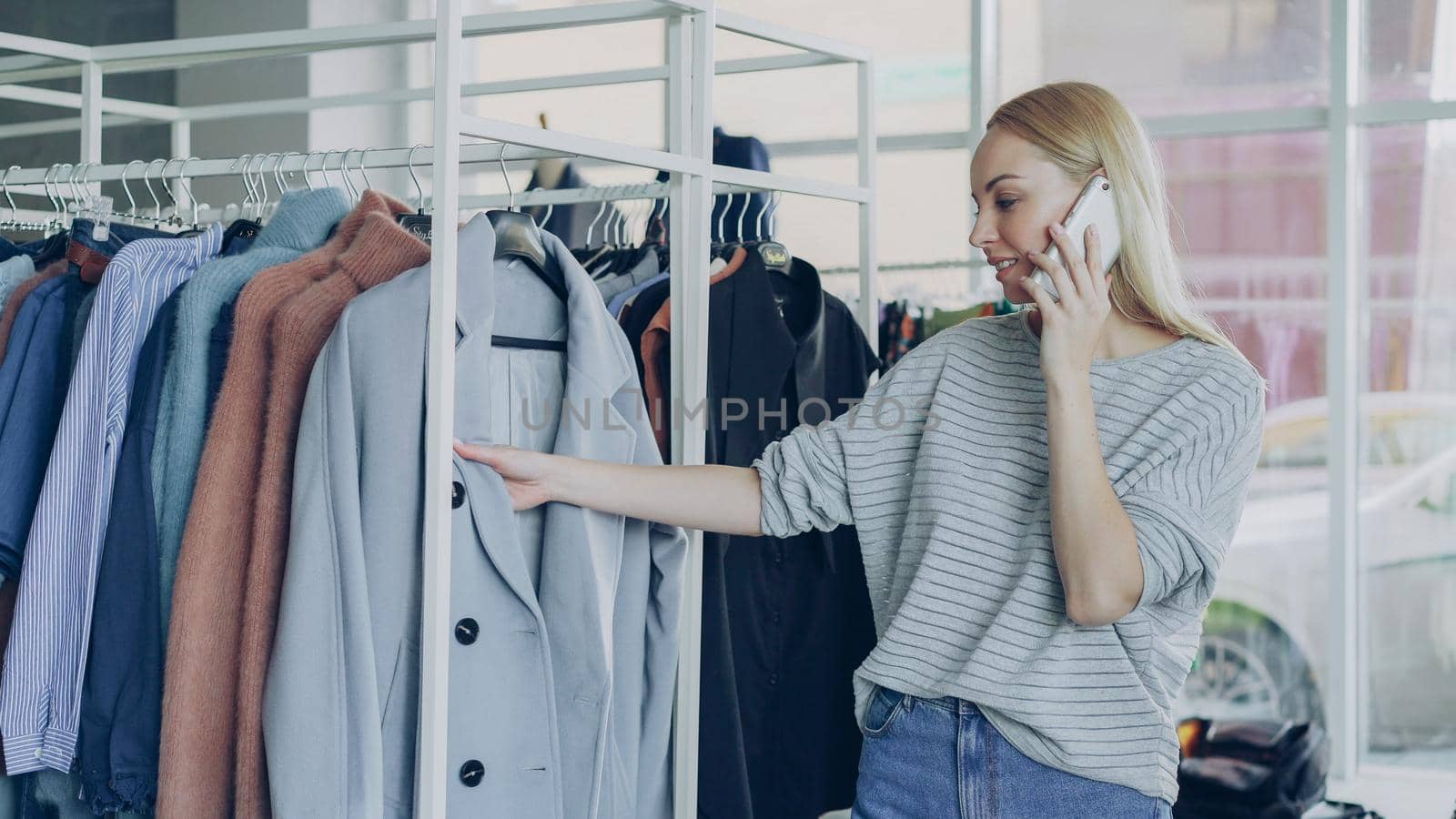 Cute blond girl is chatting on smart phone in boutique, touching clothes on hangers and smiling. Lots of fashionable clothes and large window in background. by silverkblack