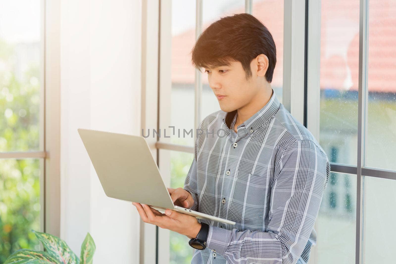 businessman smiling and working on a laptop computer stand near window in office by Sorapop