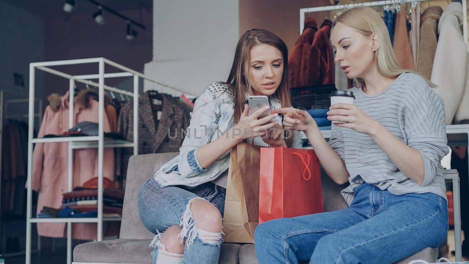 Attractive young ladies sitting on chairs in a clothing store with coffee and shopping bags, checking smartphone and talking. Shelves and hangers with colourful clothes in background