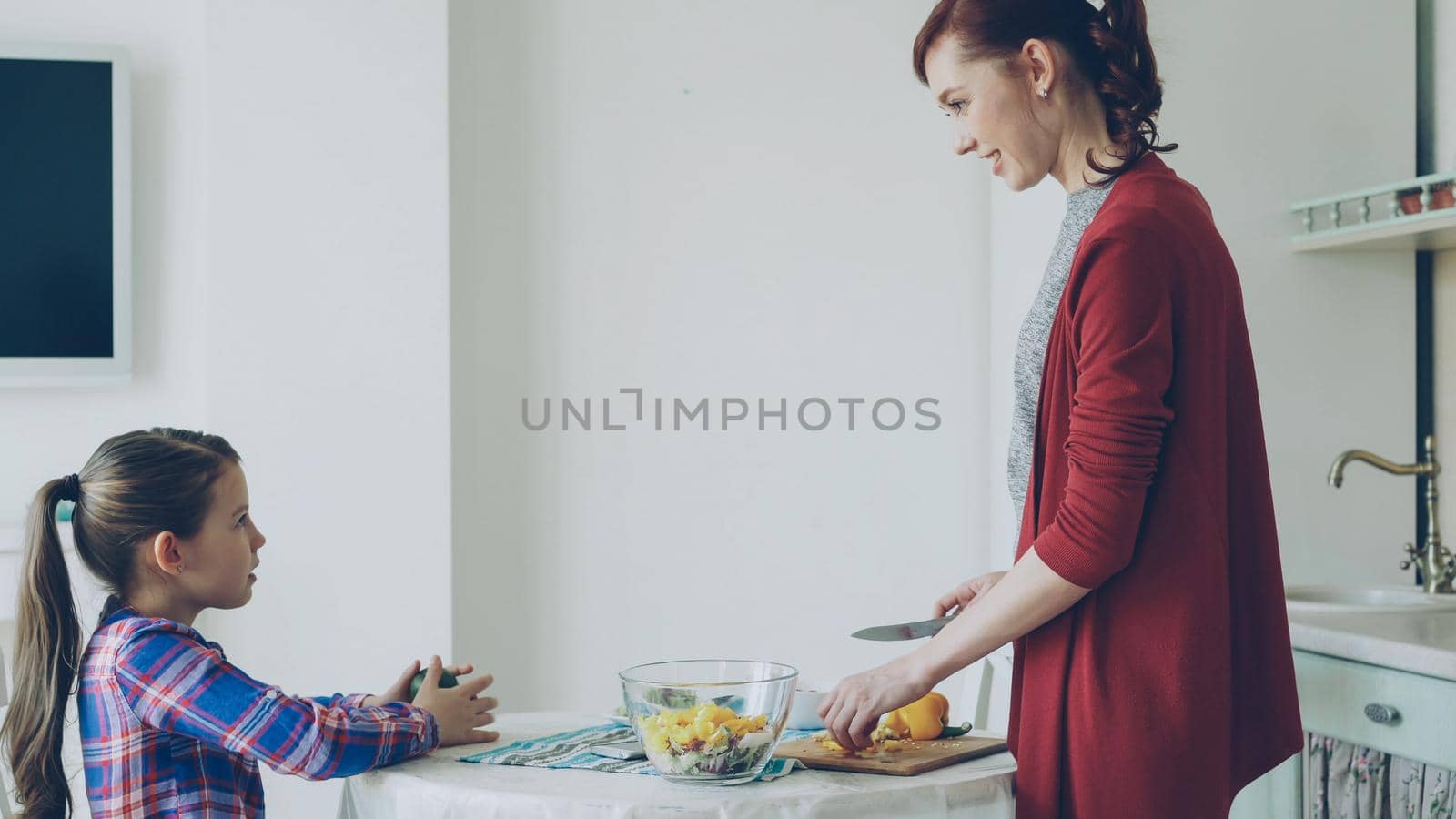 Cute funny daughter and cheerful mother talking in modern kitchen during cooking. Mom feeding her girl with piece of vegetable smiling. Family, food, home and people concept