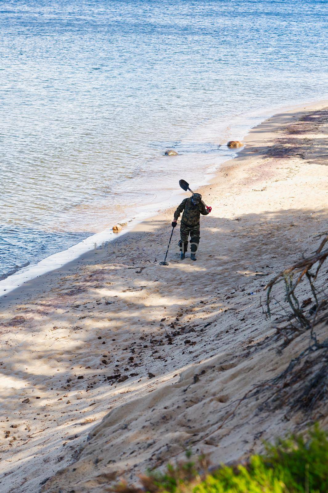 A man with a metal detector walks at the water's edge of a sandy beach. by PhotoTime