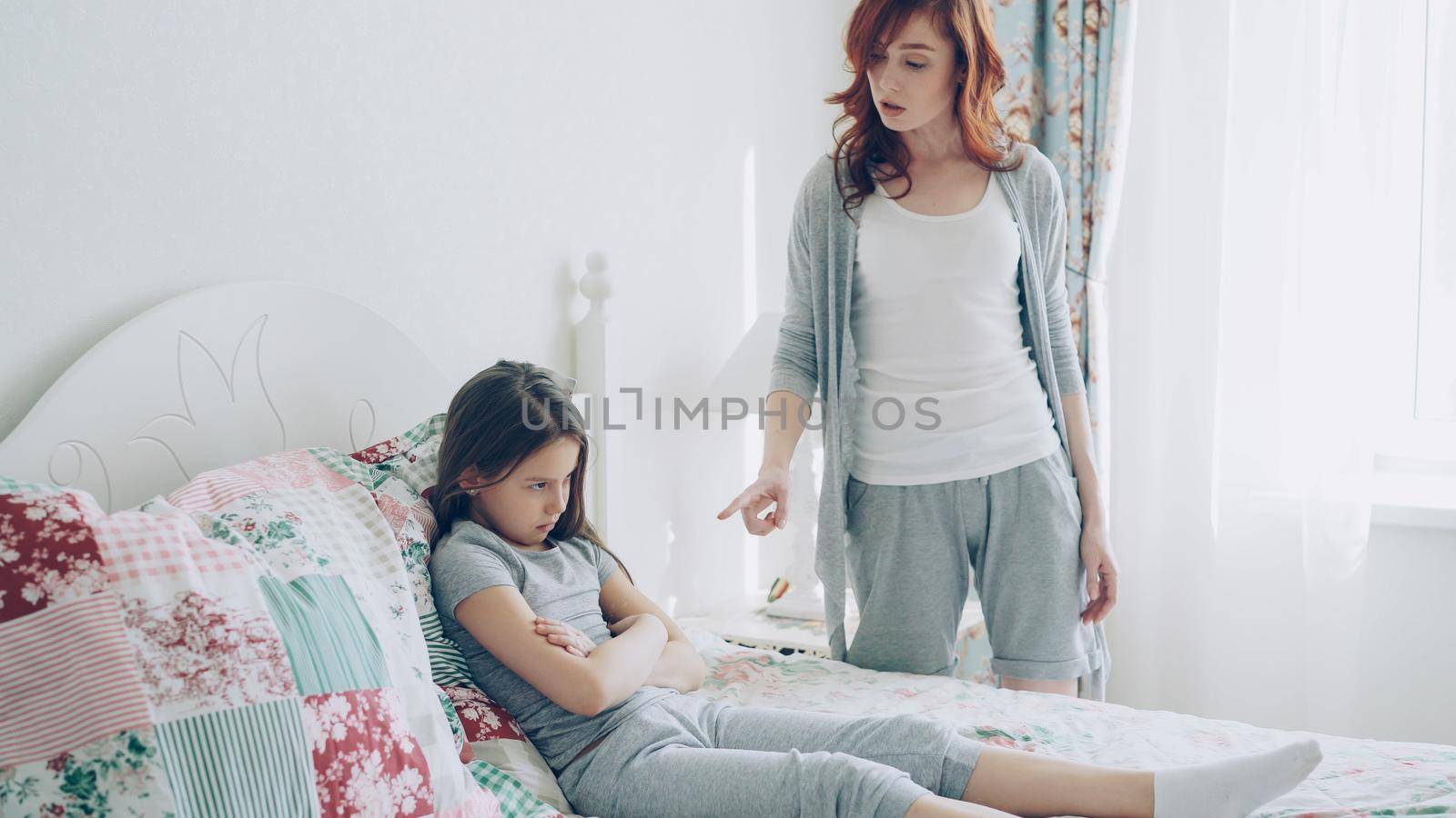 Angry mother argue scolding her upset little daughter lying on bed in bright bedroom at home by silverkblack