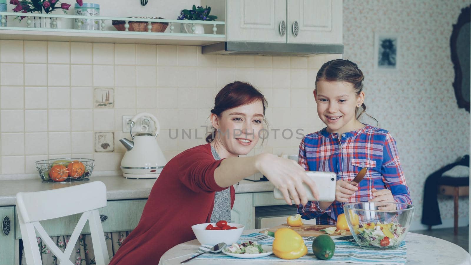 Smiling mother making selfie photo together with young cute daughter cooking breakfast at home in kitchen. Family, cook, and people concept by silverkblack