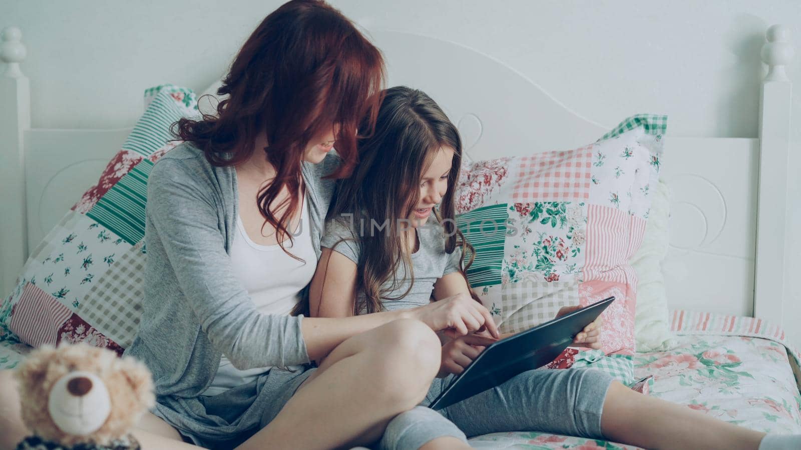 Smiling cute girl and her young mother laughing and using digital tablet while sitting on bed at home in the morning. Technology, home and family concept