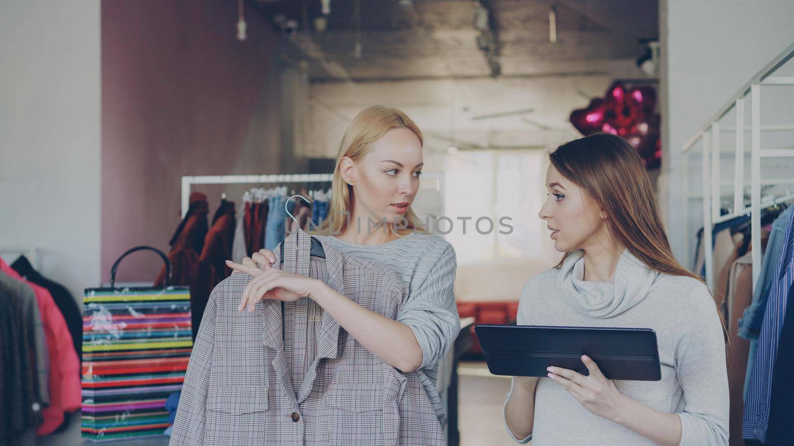 Young business owner is using tablet while standing in her clothing store. Her assistant is coming with garment and asking advice. Businesswoman is showing her where to place it. by silverkblack