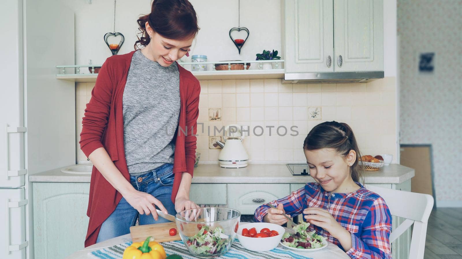 Happy mother and cheerful daughter cook salad together in the kitchen at home cutting vegetables and talking. Family, cooking, and people concept