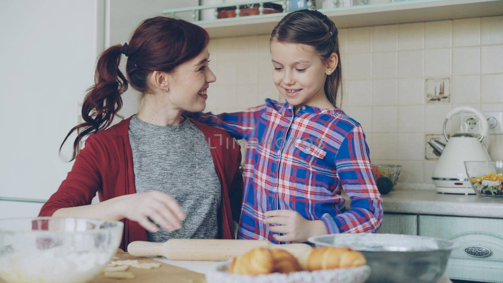 Cheerful mother cooking in the kitchen while cute daughter coming and embracing mom in the morning. Family, food, home and people concept by silverkblack