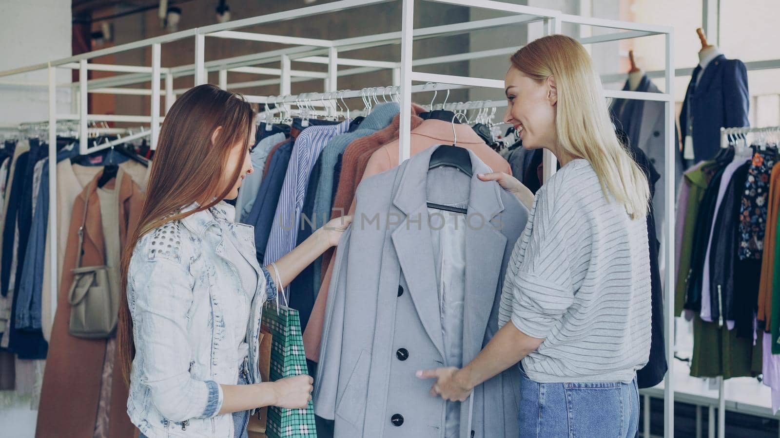 Young woman is helping her friend to find coat. She is bringing her new model, telling about it and showing its advantages. Girls are talking and smiling. by silverkblack