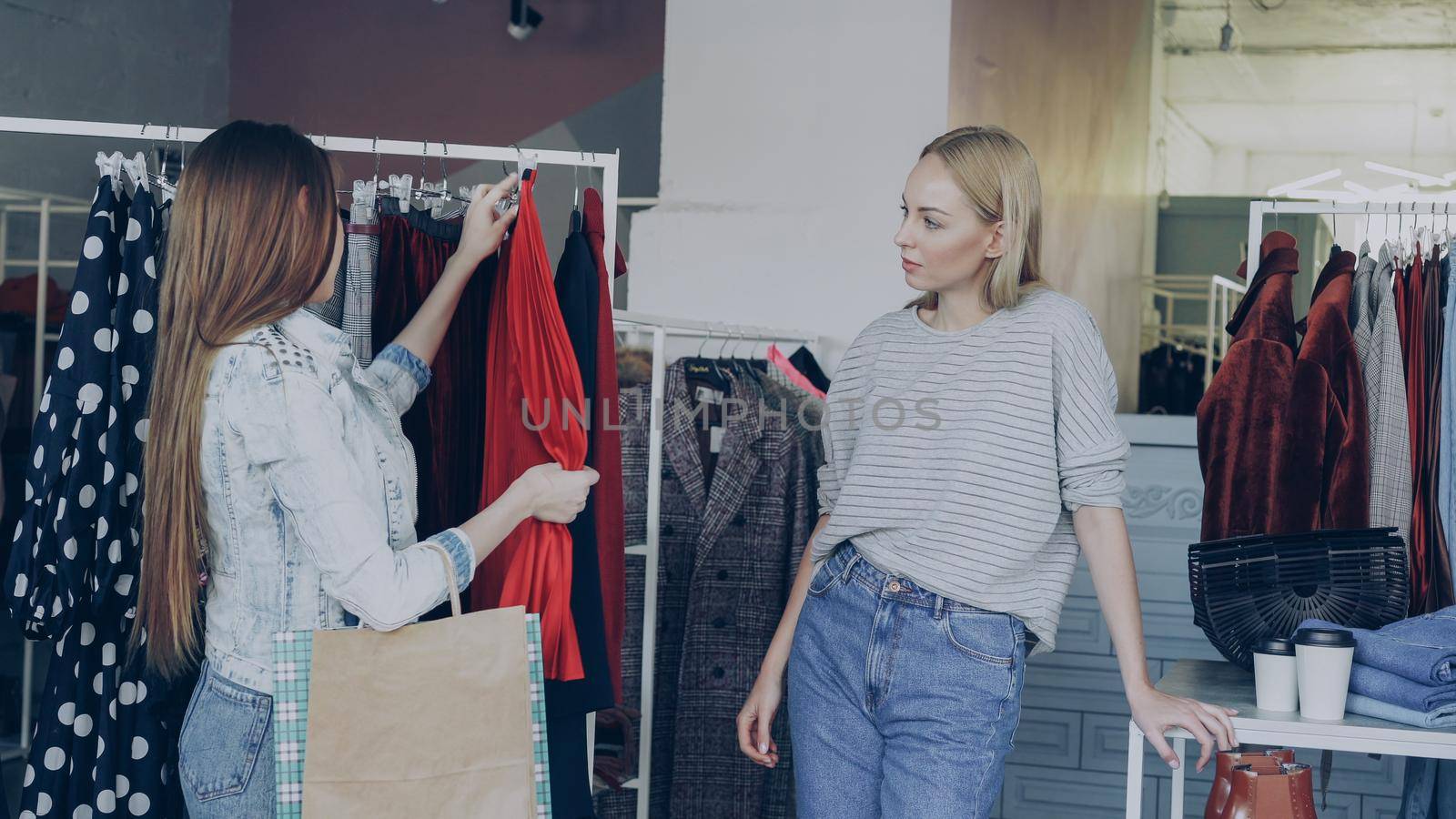 Female friends are shopping together. They are standing beside rails with stylish women's clothes, taking pleated skirt, looking at it carefully and fitting it while chatting and smiling