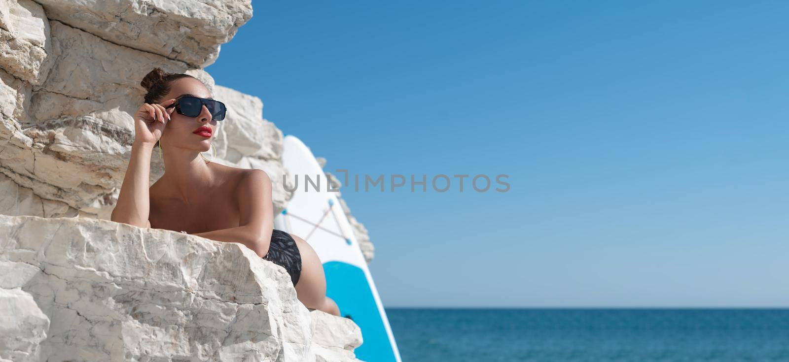 sexy young girl in a swimsuit on a sup board by the sea on the rocks in stylish sunglasses