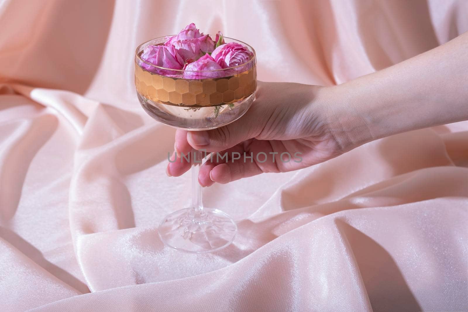 Female hand with beverage and flowers in glass on folded fabric background. Creative holiday concept