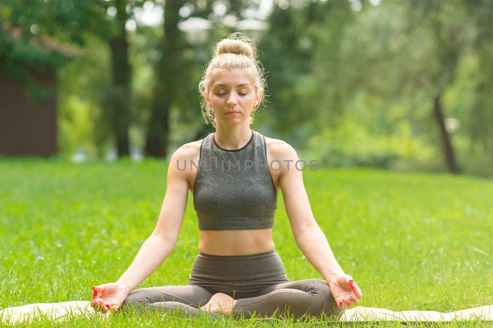 A slender woman in a gray top and leggings, sitting on mat on the green grass in summer, in the park, meditation. Copy space