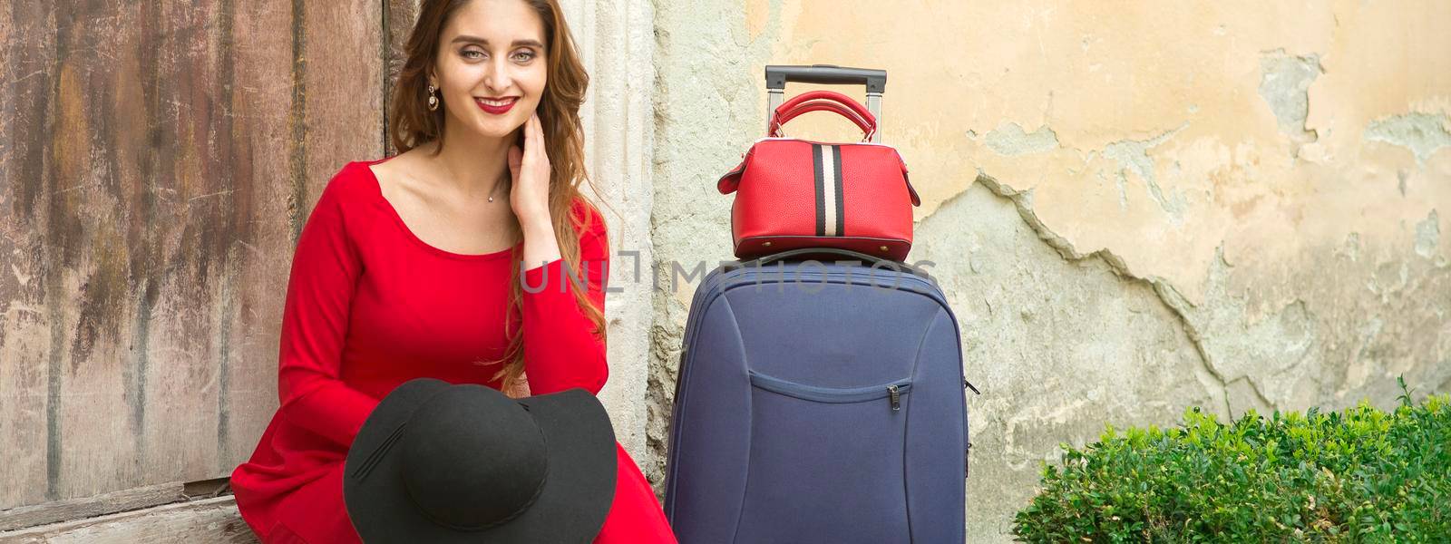 A young caucasian woman is sitting on the threshold of an old house in a red long dress with suitcase looking at the camera outdoors