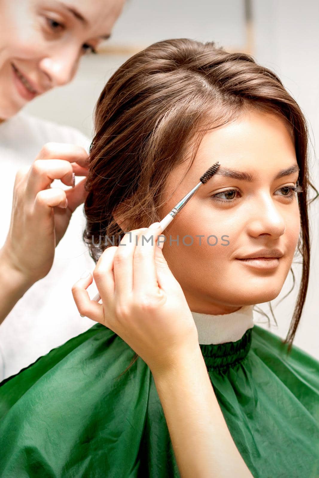 Makeup artist combing eyebrows and hairstylist preparing hairstyle for beautiful young caucasian bride in a beauty salon. Tandem of two beauty masters