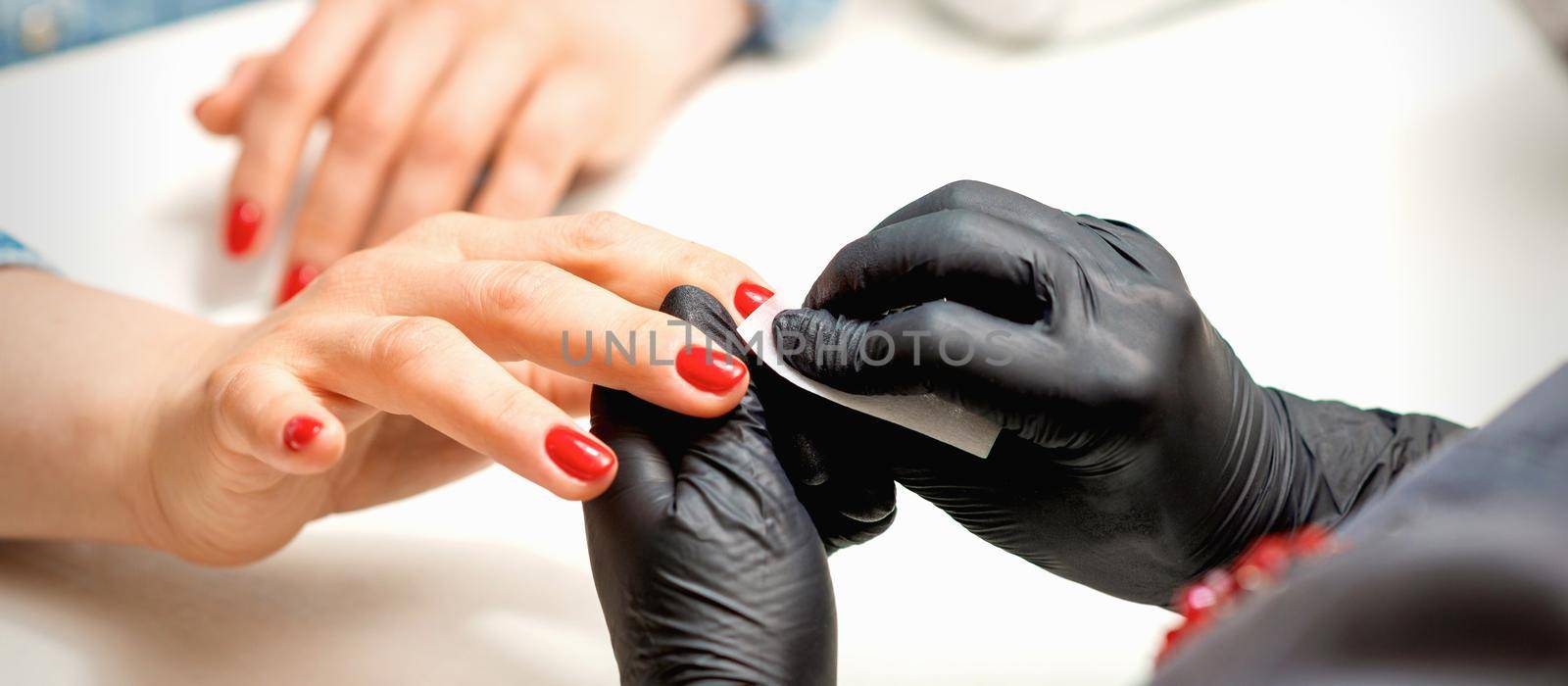 Closeup of manicure master in black gloves wiping the female red nails with a paper napkin in the beauty salon